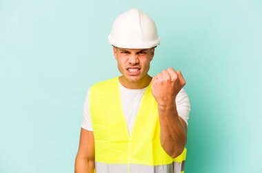 Mavi arka planda izole edilmiş genç işçi kameraya yumruk, agresif yüz ifadesi gösteriyor..