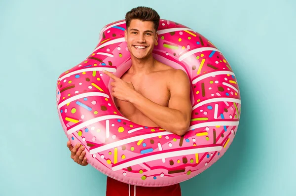 Mavi arka planda izole edilmiş şişme bir çörek tutan genç beyaz adam gülümsüyor ve boş alanda bir şey gösteriyor..