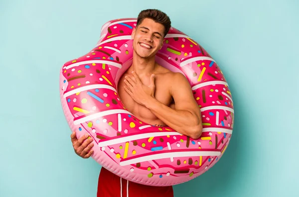 Mavi arka planda izole edilmiş şişme bir çörek tutan genç beyaz adam kahkahalar atıyor..