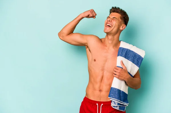 Młody Biały Mężczyzna Trzymając Ręcznik Plażowy Odizolowany Niebieskim Tle Podnosząc — Zdjęcie stockowe
