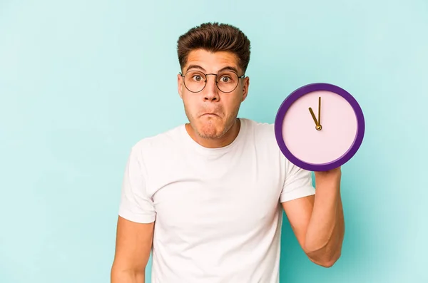 Young Caucasian Man Holding Clock Isolated Blue Background Shrugs Shoulders — Stok fotoğraf