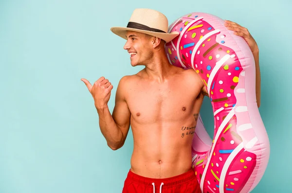 Kafkasyalı genç bir adam mavi arka plan noktalarında izole edilmiş şişme bir çörek tutuyor. Baş parmak uzakta, güler yüzlü ve kaygısız..