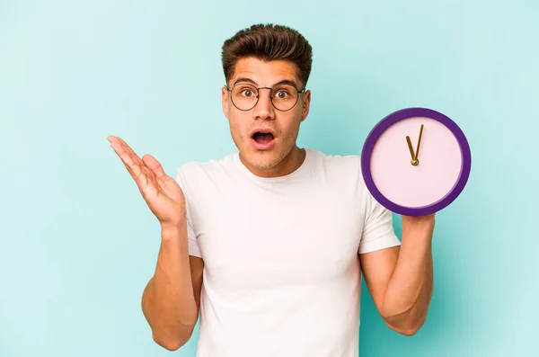 Joven Hombre Caucásico Sosteniendo Reloj Aislado Sobre Fondo Azul Sorprendido — Foto de Stock