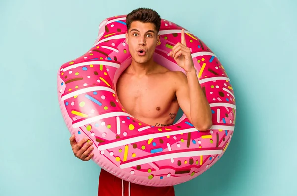 Mavi arka planda izole edilmiş şişirilebilir donut tutan genç beyaz bir adam harika bir fikre, yaratıcılık kavramına sahip..