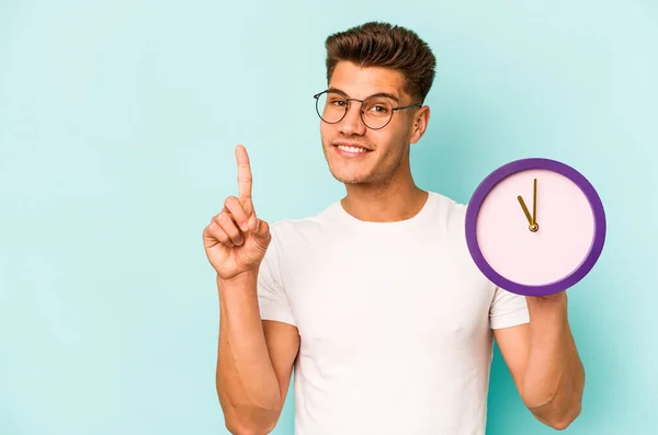 Young Caucasian Man Holding Clock Isolated Blue Background Showing Number — Φωτογραφία Αρχείου