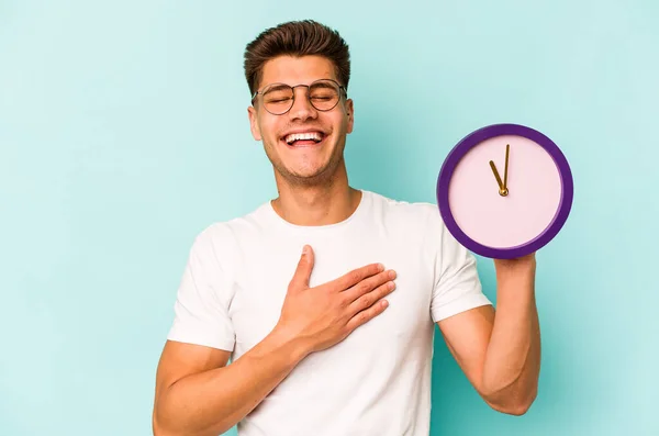 Young Caucasian Man Holding Clock Isolated Blue Background Laughs Out — стоковое фото