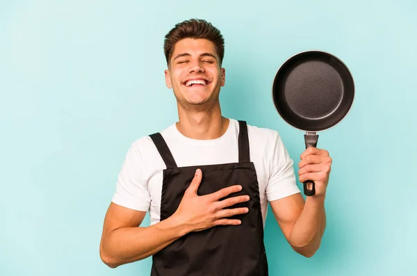 青い背景に隔離されたフライパンを保持若い白人調理師は大声で手を保つ胸に笑う — ストック写真
