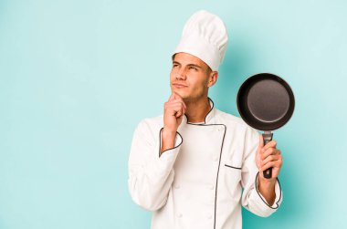 Kafkasyalı genç aşçı elinde uçan tavayla mavi arka planda izole edilmiş şüpheli ve şüpheci bir ifadeyle yana bakıyor..