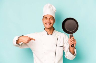 Kafkasyalı genç aşçı elinde uçan tavayla mavi arka planda izole edilmiş gömlek fotokopi alanını işaret eden gururlu ve kendinden emin.