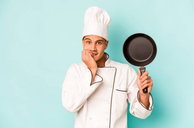 Kafkasyalı genç aşçı elinde uçan tavayla mavi arka planda izole edilmiş tırnak yiyor, gergin ve endişeli..