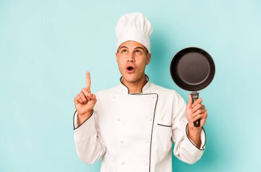 Kafkasyalı genç aşçı elinde uçan tavayla mavi arka planda izole edilmiş ağzı açık bir şekilde yukarı işaret ediyor..