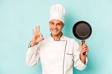 Kafkasyalı genç aşçı elinde uçan tavayla mavi arka planda izole edilmiş neşeli ve kendinden emin bir şekilde iyi niyet gösteriyor..