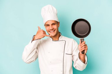 Kafkasyalı genç aşçı elinde uçan tavayla mavi arka planda izole edilmiş bir şekilde parmaklarıyla bir cep telefonu hareketi gösteriyor..