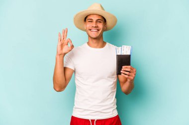 Pasaportu elinde tutan beyaz bir genç adam mavi arka planda izole edilmiş neşeli ve kendinden emin bir şekilde iyi niyet gösteriyor..