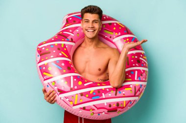 Mavi arka planda izole edilmiş şişme donut tutan genç beyaz bir adam bir avuç içinde fotokopi alanı ve belinde başka bir el tutuyor..