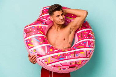 Kafkasyalı genç bir adam mavi arka planda izole edilmiş şişirilebilir bir çörek tutuyor. Kafasının arkasına dokunuyor, düşünüyor ve bir seçim yapıyor..