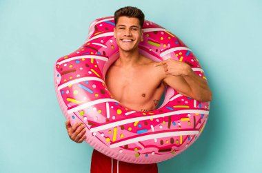 Mavi arka planda izole edilmiş şişme donut tutan genç beyaz adam gömlek fotokopi alanını işaret ediyor, gururlu ve kendinden emin.