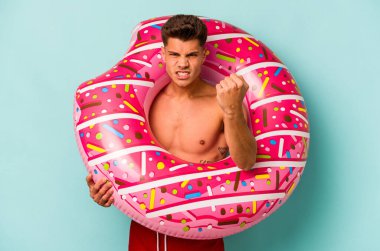 Mavi arka planda izole edilmiş şişme donut tutan genç beyaz bir adam kameraya yumruk gösteriyor, agresif bir yüz ifadesi..