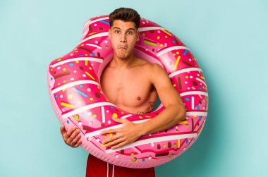 Mavi arka planda izole edilmiş şişme bir çörek tutan genç beyaz adam omuzlarını silker ve gözleri açık kalır..