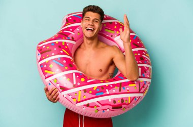 Mavi arka planda izole edilmiş şişme bir çörek tutan genç beyaz adam hoş bir sürpriz alıyor, heyecanlı ve el kaldırıyor..