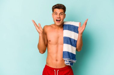 Elinde plaj havlusu tutan genç beyaz adam mavi arka planda izole edilmiş, şaşırmış ve şok olmuş..