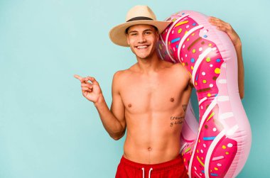 Mavi arka planda izole edilmiş şişme bir çörek tutan genç beyaz adam gülümsüyor ve boş alanda bir şey gösteriyor..