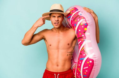 Mavi arka planda izole edilmiş şişme donut tutan genç beyaz adam işaret parmağıyla hayal kırıklığı jesti gösteriyor..