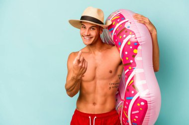 Mavi arka planda izole edilmiş şişme donut tutan genç beyaz bir adam sanki seni davet ediyormuş gibi seni işaret ediyor..