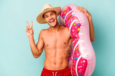 Mavi arka planda izole edilmiş şişme bir çörek tutan genç beyaz adam neşeli ve tasasız parmaklarıyla barış sembolü gösteriyor..