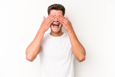 Beyaz arka planda soyutlanmış beyaz tenli genç bir adam elleriyle gözlerini kapatıyor, geniş bir gülümsemeyle bir sürpriz bekliyor..