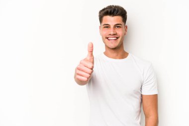 Beyaz arka planda izole edilmiş genç beyaz adam gülümsüyor ve başparmağını kaldırıyor.