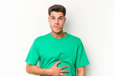 Beyaz arka planda izole edilmiş genç beyaz adam karnına dokunuyor, nazikçe gülümsüyor, yemek yiyor ve tatmin oluyor..