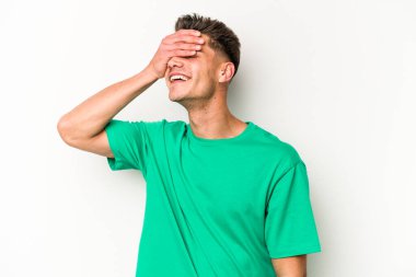 Beyaz arka planda soyutlanmış beyaz tenli genç bir adam elleriyle gözlerini kapatıyor, geniş bir gülümsemeyle bir sürpriz bekliyor..