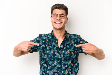 Beyaz arka planda yaşayan beyaz tenli genç bir adam gömlek fotokopi alanını gösteriyor, gururlu ve kendinden emin.