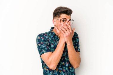 Beyaz arka planda izole edilmiş genç beyaz bir adam parmaklarının arasından korkmuş ve gergin bir şekilde göz kırpıyor..