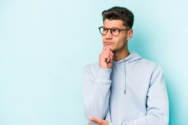 Mavi arka planda izole edilmiş genç beyaz adam şüpheli ve şüpheci bir ifadeyle yana bakıyor..