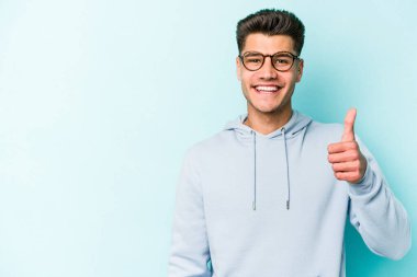 Mavi arka planda izole edilmiş genç beyaz adam gülümsüyor ve başparmağını kaldırıyor.