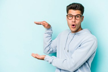 Mavi arka planda izole genç beyaz adam şok ve hayret içinde elleri arasında bir kopya boşluk tutuyor.