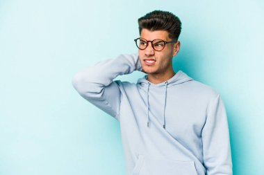 Kafkasyalı genç adam mavi arka planda izole edilmiş kafasının arkasına dokunuyor, düşünüyor ve bir seçim yapıyor..