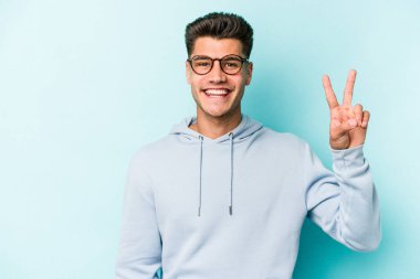 Mavi arka planda izole edilmiş genç beyaz adam zafer işareti gösteriyor ve gülümsüyor..