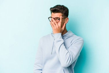 Mavi arka planda izole edilmiş genç beyaz adam kameraya parmaklarıyla göz kırpıyor, yüzünü kapatmaktan utanıyor..