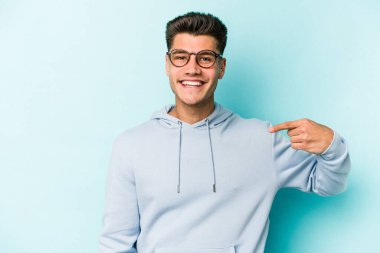 Mavi arka planda izole edilmiş genç beyaz adam bir gömlek fotokopi alanını işaret ediyor, gururlu ve kendinden emin.