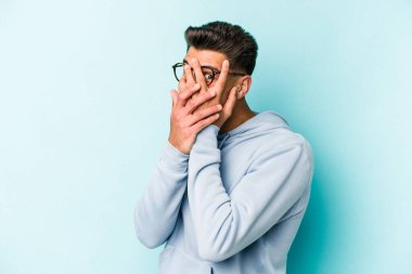 Kafkasyalı genç adam mavi arka planda izole edilmiş. Korkmuş ve gergin parmaklarıyla göz kırp..