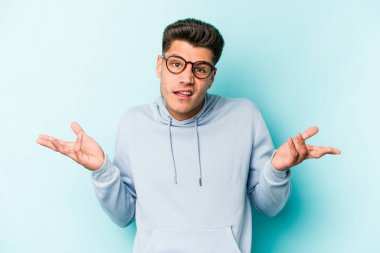 Kafkasyalı genç adam mavi arka planda izole edilmiş sorgulama jestinde şüphe ediyor ve omuz silkiyor..