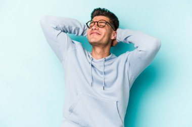 Kafkasyalı genç adam mavi arka planda izole edilmiş. Kendine güveniyor, elleri başının arkasında..