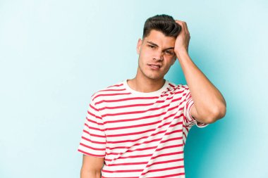 Kafkasyalı genç adam mavi arka planda izole edilmiş. Yorgun ve uykulu. Elini başının üstünde tutuyor..