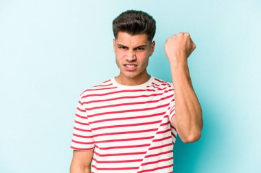 Mavi arka planda izole edilmiş genç beyaz adam yumruğunu kameraya gösteriyor, agresif yüz ifadesi var..