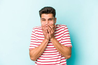 Mavi arka planda izole edilmiş genç beyaz bir adam bir şeye gülüyor, ağızlarını ellerle kapatıyor..