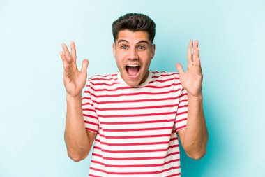 Mavi arka planda izole edilmiş genç beyaz bir adam zaferi ya da başarıyı kutluyor, şaşırıyor ve şok oluyor..