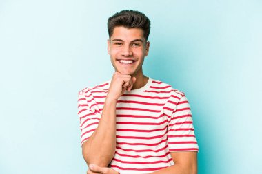 Mavi arka planda izole edilmiş genç beyaz adam gülümsüyor ve kendinden emin, elleriyle çenesine dokunuyor..